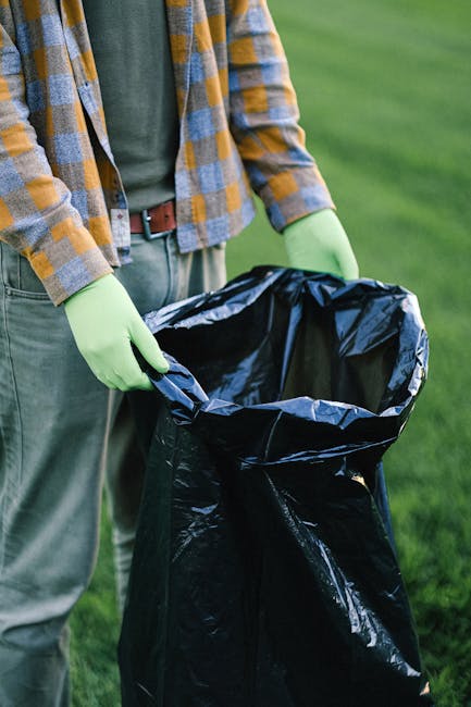 A person standing outdoors on a grassy area, wearing a yellow and blue checkered shirt, grey trousers, and lime green gloves, holding open a large black plastic rubbish bag with both hands. The bag appears to be empty or in the process of being filled with waste, with its top rolled over slightly. The background shows a blurred green landscape, indicating a park or garden environment. The scene suggests an on-site waste collection or rubbish removal activity, typical of private rubbish disposal services such as those offered by Waste Collection Brixton, which provides alternative waste handling solutions beyond standard municipal rubbish collection.
