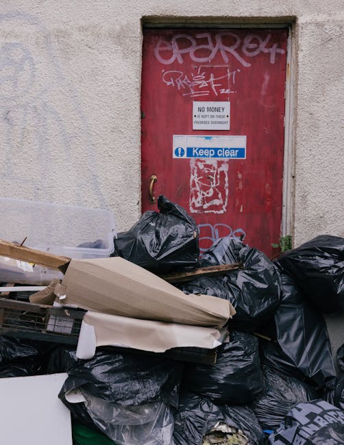 A pile of black garbage bags and discarded packaging materials, including flattened cardboard boxes and pieces of white foam, are stacked against a weathered exterior wall beneath a small, rectangular red door with graffiti tags and multiple stickers. One sticker displays the phrase 'Keep clear' in white text on a blue background, and another sign reads 'No Money - No Dock on Time - Payment Overdue.' The scene appears to be an urban alley or side street, with the waste collection situated near a building wall with a rough, textured surface. The overall atmosphere suggests a private waste disposal point or on-site rubbish clearance, with waste temporarily accumulated for collection, reflecting the practical aspect of alternative rubbish removal services like those offered by Waste Collection Brixton. The lighting is natural, casting minimal shadows, highlighting the cluttered and unmanaged state of the rubbish pile and the slightly worn condition of the surrounding environment.