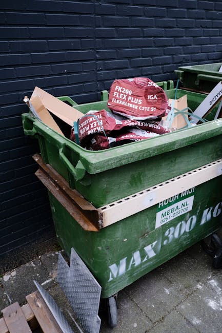 A large green waste bin filled with red and black bags of garden or building debris, placed outdoors beside a dark brick wall and a concrete pavement. The bin is supported by a white plastic frame resting on a wooden pallet, and the contents include bags labeled 'FLEXLEEBER CZTE S1' and similar. Adjacent to the bin, there are additional green waste containers, suggesting a setting for waste collection or disposal. The environment appears to be an alleyway or back area, typical for onsite rubbish removal services such as those offered by Waste Collection Brixton, providing an alternative to standard council collection for garden or building waste.