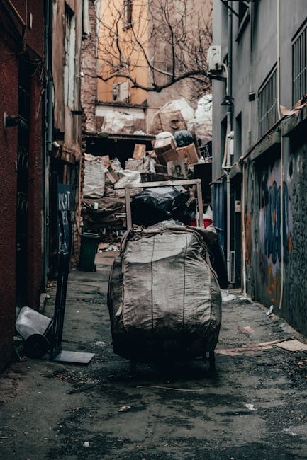 A narrow urban alleyway with a grey, weathered pavement showing signs of dirt and debris, flanked on both sides by tall buildings with graffiti and peeling paint. In the foreground, a large, grey, woven polypropylene sack filled with waste materials is positioned in the center, partially blocking the pathway. Behind it, a utility shopping trolley with a metal frame and fabric bag is loaded with miscellaneous rubbish. Further down the alley, a substantial pile of assorted discarded items, including cardboard boxes, black plastic bags, and loose debris, rests against a makeshift wooden barrier at the back of the alley, which is cluttered with waste. The surroundings suggest an environment used for private waste disposal, and the scene exemplifies a typical site for independent rubbish collection, managed by Waste Collection Brixton, highlighting the challenges of limited access in narrow spaces and the need for specialized disposal services in densely built-up areas. Overhead, leafless tree branches extend into the view, and the lighting is natural but subdued, creating a somewhat gloomy atmosphere associated with waste removal and clutter accumulation.
