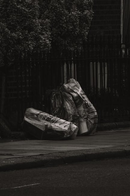 A group of black plastic garbage bags filled with waste are placed on the street pavement, positioned near a black metal fence with vertical bars and a lattice design. The bags appear to contain household or garden refuse, with some partially crumpled and bulging, showing the irregular shapes of the waste inside. One bag is leaning against the fence, while others are stacked slightly to the side, creating an uneven pile. The environment suggests an outdoor urban setting, with a large tree or shrub partially visible in the upper left corner, and the scene is illuminated by natural daylight, casting soft shadows on the ground. This scene exemplifies alternative waste handling or private rubbish collection, possibly managed by Waste Collection Brixton, indicated by the proximity to the fence and pavement typically associated with independent rubbish removal services in the area. The overall atmosphere is neutral and quiet, with no visible vehicles or people, focused solely on the waste bags ready for collection or disposal.