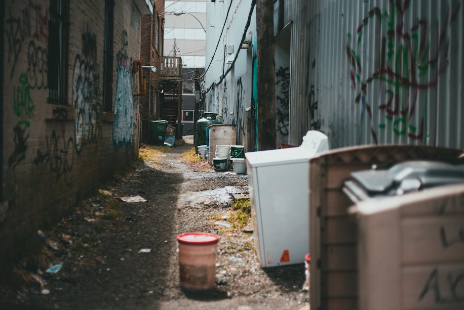 A narrow urban alleyway with a grey, weathered pavement showing signs of dirt and debris, flanked on both sides by tall buildings with graffiti and peeling paint. In the foreground, a large, grey, woven polypropylene sack filled with waste materials is positioned in the center, partially blocking the pathway. Behind it, a utility shopping trolley with a metal frame and fabric bag is loaded with miscellaneous rubbish. Further down the alley, a substantial pile of assorted discarded items, including cardboard boxes, black plastic bags, and loose debris, rests against a makeshift wooden barrier at the back of the alley, which is cluttered with waste. The surroundings suggest an environment used for private waste disposal, and the scene exemplifies a typical site for independent rubbish collection, managed by Waste Collection Brixton, highlighting the challenges of limited access in narrow spaces and the need for specialized disposal services in densely built-up areas. Overhead, leafless tree branches extend into the view, and the lighting is natural but subdued, creating a somewhat gloomy atmosphere associated with waste removal and clutter accumulation.