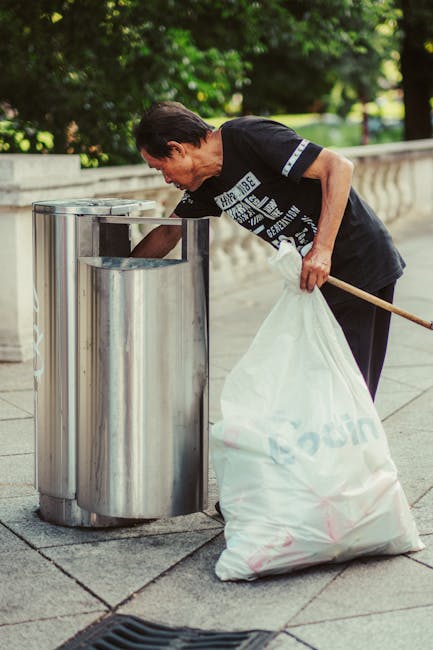 A middle-aged woman with dark hair pulled back, dressed in a black T-shirt with white and blue graphics and black trousers, is seen disposing of rubbish into a metallic public trash bin on a paved area. She is holding a large white plastic bag filled with waste in one hand and a wooden stick or broom handle in the other. The trash bin is cylindrical, made of stainless steel, with a smooth, shiny surface and a rectangular opening at the top. The scene is outdoors, with lush green trees and bushes in the background, and a stone balustrade lining the perimeter of the walkway. Soft sunlight illuminates the scene, creating natural shadows and highlighting the textures of the metallic bin, the bag's plastic surface, and the paved ground. The visual suggests an emphasis on personal waste disposal or local waste management efforts, which could relate to private or alternative rubbish collection services like those offered by Waste Collection Brixton.