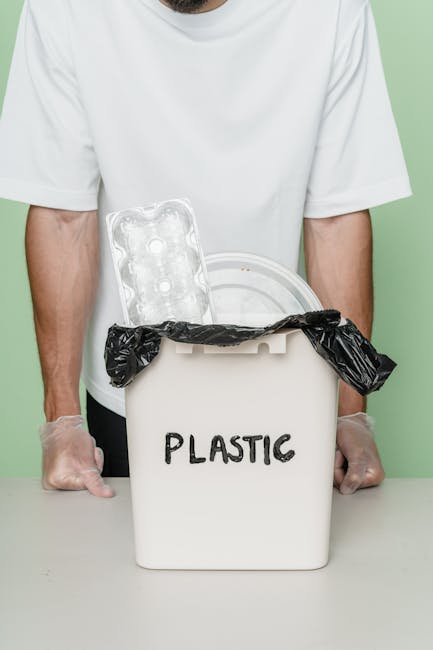 A person wearing a white t-shirt and transparent gloves holds a white plastic bin labeled 'PLASTIC' in black letters. The bin contains a black plastic rubbish bag at the top, partially open, with a clear plastic tray placed inside, leaning against the bag. The scene is set against a plain, light-colored background, emphasizing the focus on waste disposal of plastic materials, which relates to services provided by companies like Waste Collection Brixton for alternative rubbish removal and on-site clearance.