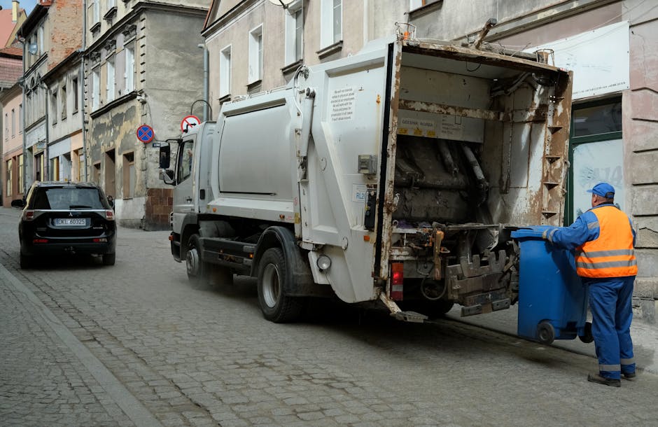 A street scene showing a waste collection truck operated by Waste Collection Brixton parked along a narrow cobblestone road in an urban environment. The truck is primarily white, with visible signs of rust and wear on the rear loading area, which is open and ready to collect rubbish. A worker wearing a high-visibility orange vest, blue overalls, and a blue cap is positioned at the rear, actively placing a blue wheeled bin into the truck's hopper. The on-site clearance process involves manual loading of waste, indicating an alternative or private waste collection method rather than standard council rubbish disposal. In the background, residential buildings with varied stone and brick facades line the street, some with partially peeling paint and small windows. A parked black car is visible further down the street, and a no-parking sign is attached to a lamppost. The lighting suggests daytime, with natural light illuminating the scene, emphasizing the gritty texture of the cobbled surface and the utilitarian design of the waste collection equipment.