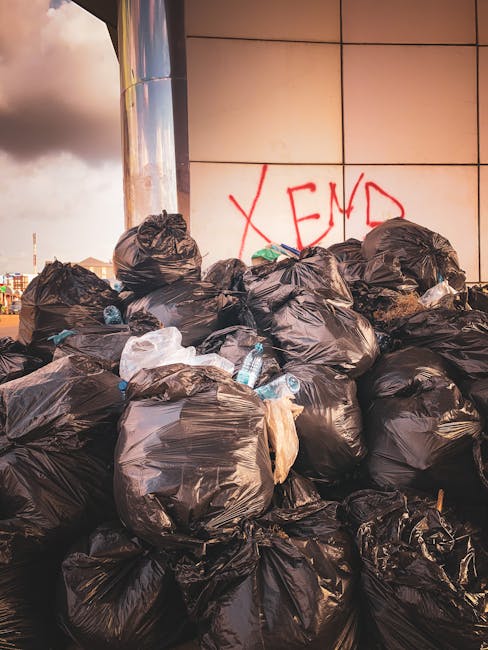 A person wearing a dark beanie, a high-visibility yellow vest, and dark clothing stands on a train station platform next to an orange hand-pulled trolley heavily loaded with various bags of rubbish, including black, grey, and orange plastic garbage bags, some appearing full and tied, along with a larger black container or bin. The trolley is positioned beside a modern glass-walled elevator shaft, with robotic waste collection equipment visible behind it. In the background, there are train tracks with a stationary train on the left side of the platform, and a small group of indistinct people further along the platform on the right. The platform surface is smooth and clean, made of large rectangular tiles, and the overall scene is illuminated by diffuse natural light, creating a neutral, professional atmosphere that reflects an environmental context of waste handling or alternative rubbish disposal at a busy urban train station. This scene subtly relates to private waste collection services like those provided by Waste Collection Brixton, highlighting the visual aspect of independent rubbish removal in public or semi-public transport hubs.